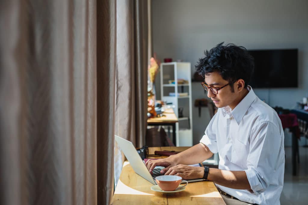 A man working from a laptop on a wooden desk with a cup of team.